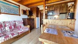 A cozy cabin interior with a wooden dining table, a sofa with a patterned cover, a kitchen area with wooden cabinets and appliances, and a framed picture on the wall.