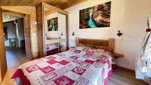 A bedroom with a wooden ceiling, a large bed with a patterned quilt, a painting of a river on the wall, and a mirror cabinet.