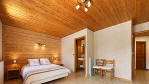 Un dormitorio con techo de madera, una cama, una mesita de noche, una silla y un baño visible a través de una puerta abierta.