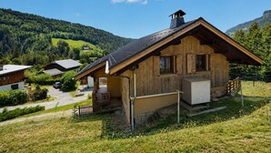 Una cabaña de madera con techo inclinado, un porche y un garaje, rodeada de un exuberante paisaje verde y montañas.
