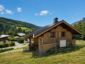 Un chalet en bois doté d’un toit à pente, d’un porche et d’un garage, entouré d’un paysage verdoyant et luxuriant ainsi que de montagnes.