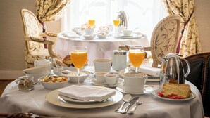 A breakfast table set with various dishes, including toast, fruit, and beverages, in a room with floral curtains and a view of the outdoors. - Aux Charmes de Maintenon - Adults Only (Maintenon)