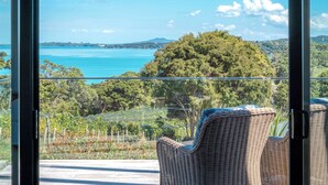A balcony with wicker chairs, a glass railing, and a view of a blue sea and green hills. - Te Whau Retreat - Guest House (Waiheke Island)