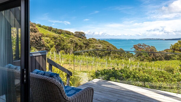 A balcony with wicker furniture, a view of a vineyard, and a distant coastline. - Te Whau Retreat - Guest House (Waiheke Island)