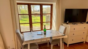 Un comedor con una mesa dispuesta para dos personas, una botella de champán y una copa. Se ve una ventana con vistas a un paisaje verde. Un televisor está montado en la pared a la derecha.