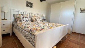 Un dormitorio con cama blanca, ropa de cama floral, una mesita de noche con lámpara y un armario.
