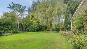 Un giardino curato con diverse piante, alberi e arbusti, un prato verdeggiante e un edificio parzialmente nascosto dalla vegetazione.