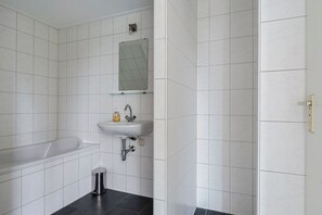 Une salle de bain avec des carreaux blancs, une baignoire, un lavabo, un miroir et une poubelle.