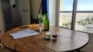 Ein runder Holztisch mit einer Flasche Bouché, zwei Gläsern und einem Smartphone. Auf dem Tisch liegen Papiere. Ein Fenster mit Balkonblick ist zu sehen.