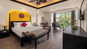 Une chambre d’hôtel moderne dotée d’un grand lit, d’un ventilateur de plafond, d’un bureau avec un panier de fruits et offrant une vue sur la verdure environnante.