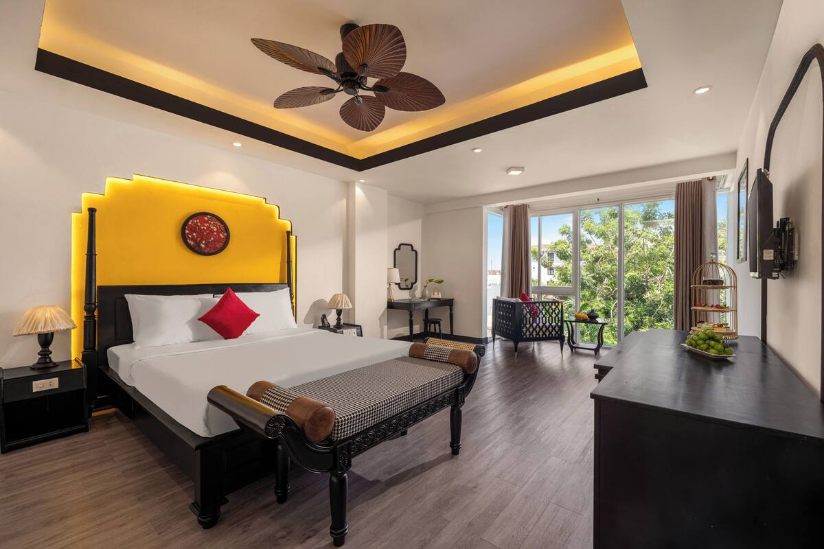 Une chambre d’hôtel moderne dotée d’un grand lit, d’un ventilateur de plafond, d’un bureau avec un panier de fruits et offrant une vue sur la verdure environnante.