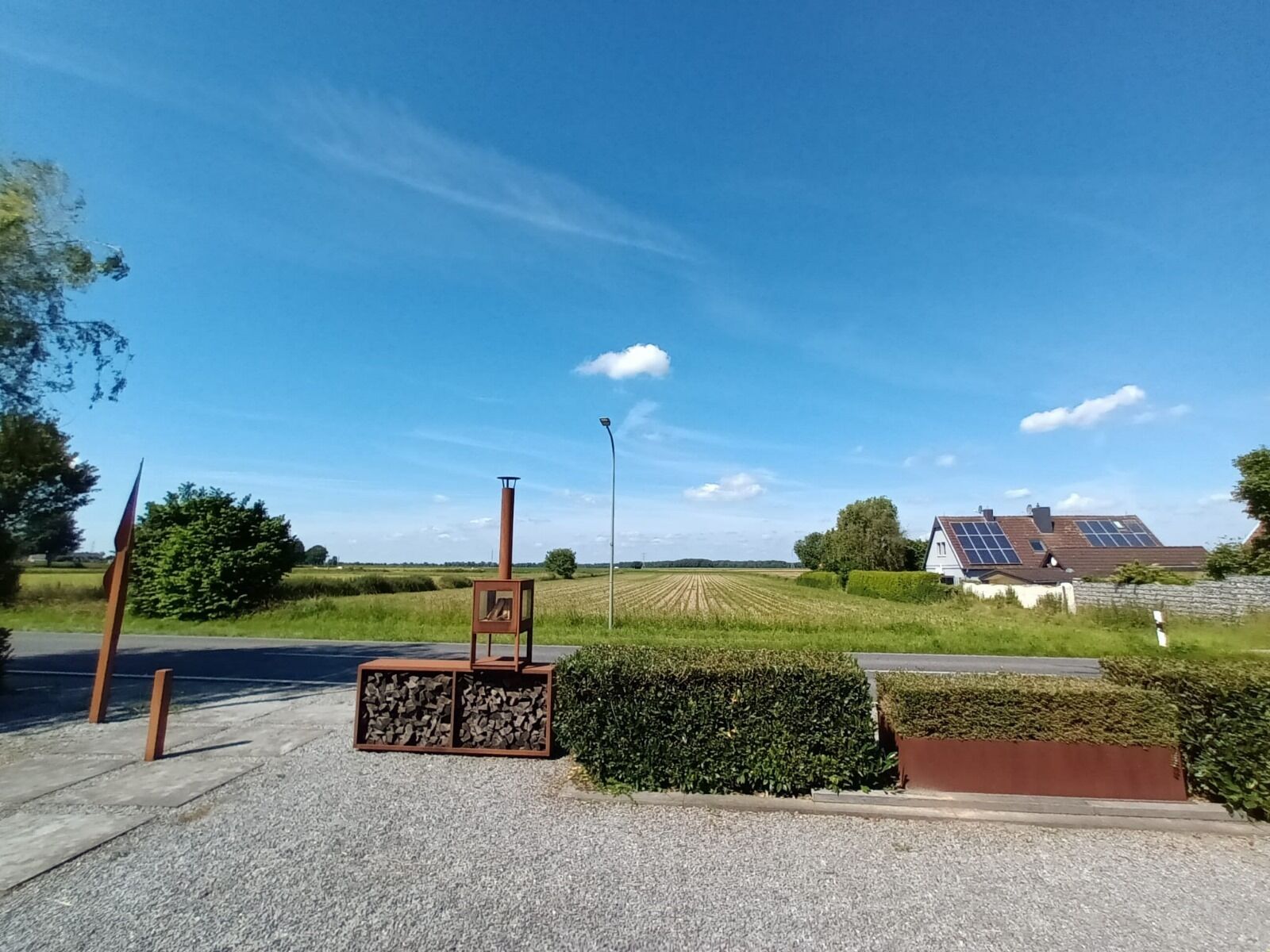 Un paysage rural avec un chemin de gravier, une sculpture en métal, une maison équipée de panneaux solaires et un champ en arrière-plan.