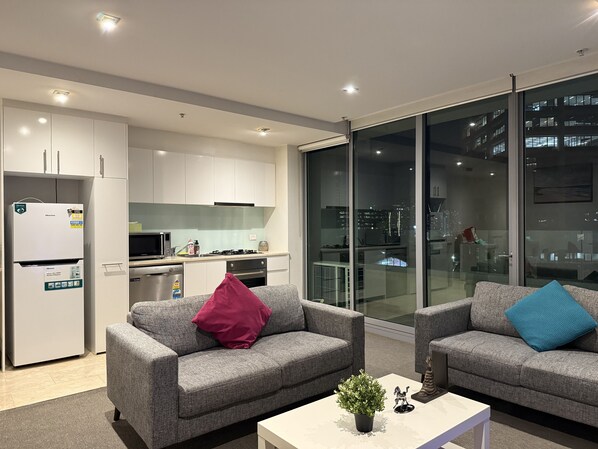 A modern living room with grey sofas, a white coffee table, and a city view through large glass windows.