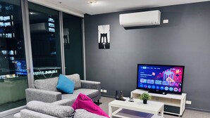 Un moderno salón con sofás grises, un televisor de pantalla plana y vistas a la ciudad a través de amplios ventanales.