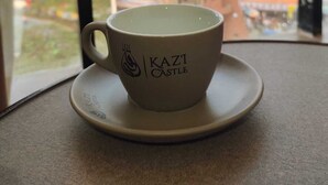 Eine Tasse mit dem Schriftzug "Kazi Castle" auf einem Untertasschen, platziert auf einem Tisch neben einem Fenster mit Blick auf ein Gebäude.