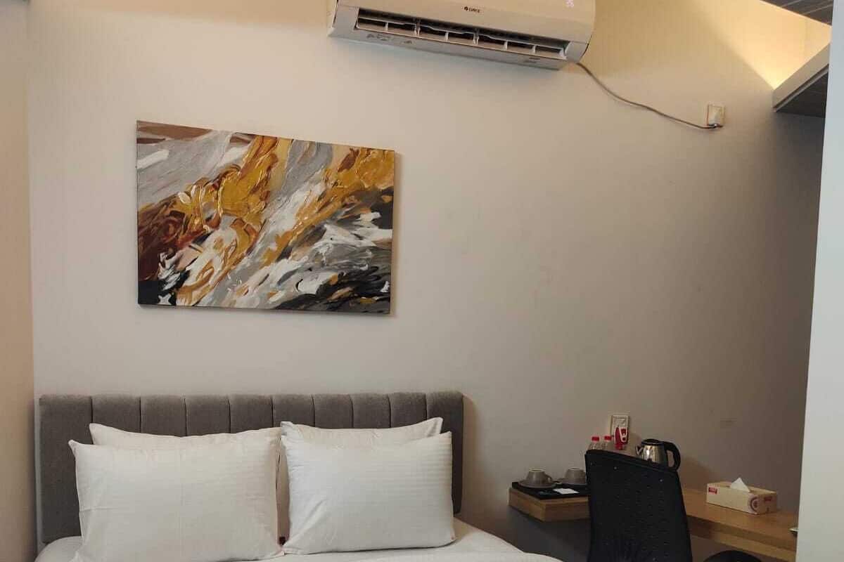 A hotel room with a bed, a gray headboard, a painting on the wall, and an air conditioning unit above the bed.
