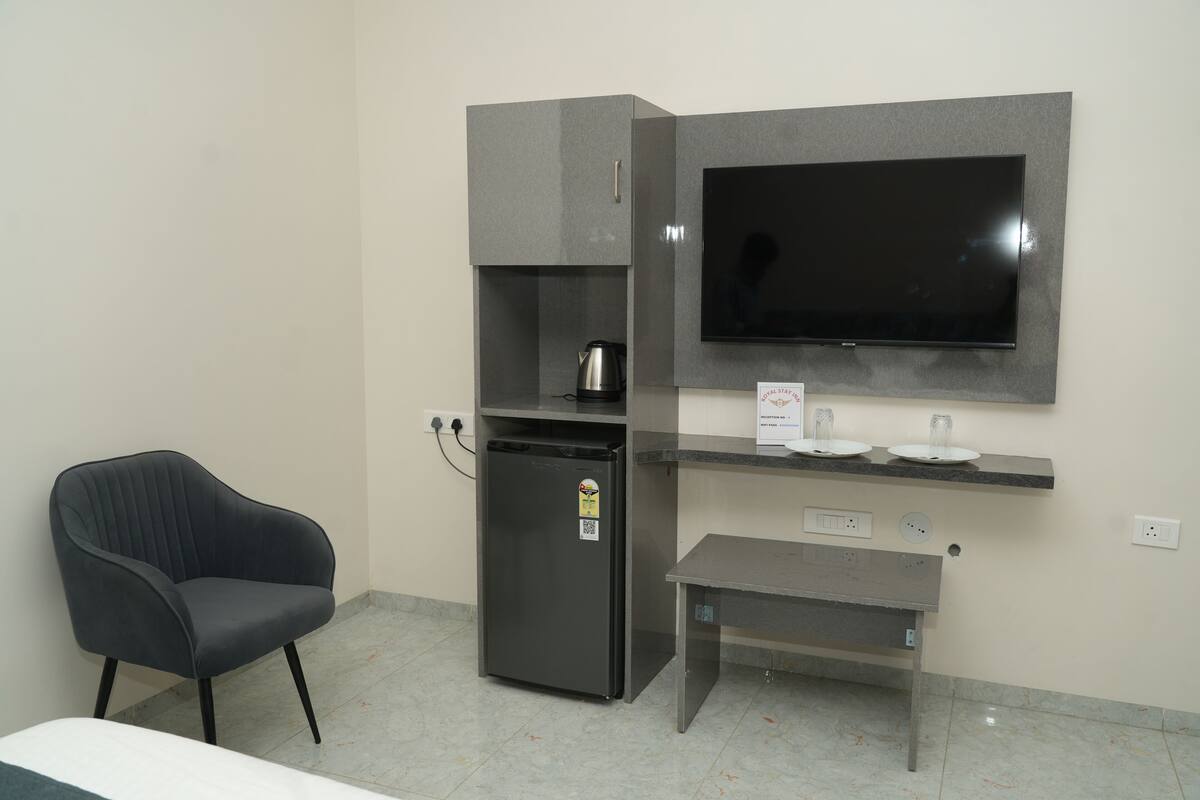 A modern room with a flat-screen TV mounted on the wall, a small refrigerator, a coffee maker, a chair, and a small table.