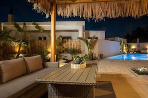 Un área junto a la piscina con una mesa de madera, muebles de mimbre y un techo de paja.