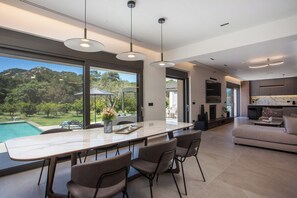 Une cuisine moderne dotée d’un grand îlot central, d’un coin repas avec des chaises, et offrant une vue sur une piscine et un espace verdoyant.