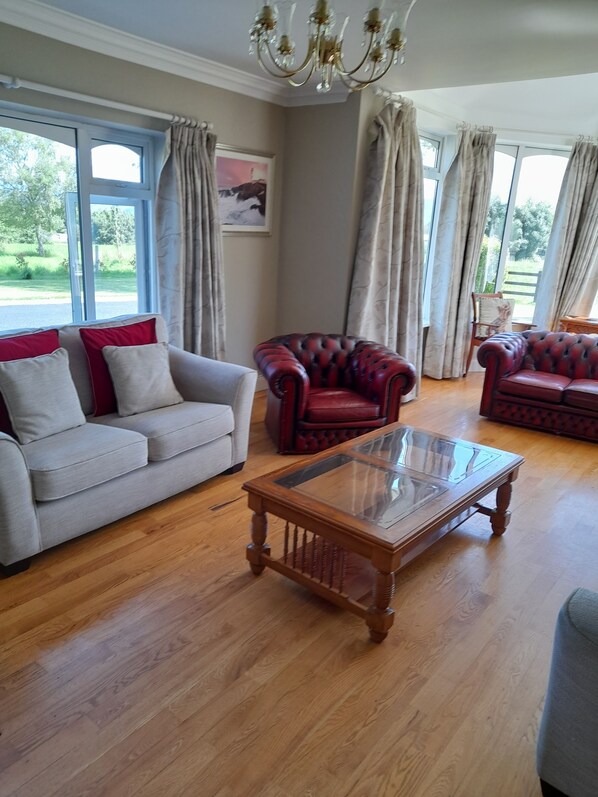 A spacious living room with a grey sofa, red leather armchair, and a glass-top coffee table. Large windows with curtains provide natural light and views of the outdoors.