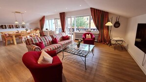 A spacious living room with a red sofa, a striped sofa, and a glass coffee table. There is a dining area with a wooden table and chairs, and a balcony with a view of the outside. - AlpenHorizont: Spacious vacation apartment with a dream view for families & friends (Garmisch-Partenkirchen)