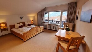 A bedroom with a wooden bed, a dining table, a chair, a view of mountains through the window, and a mounted deer head on the wall. - AlpenHorizont: Spacious vacation apartment with a dream view for families & friends (Garmisch-Partenkirchen)