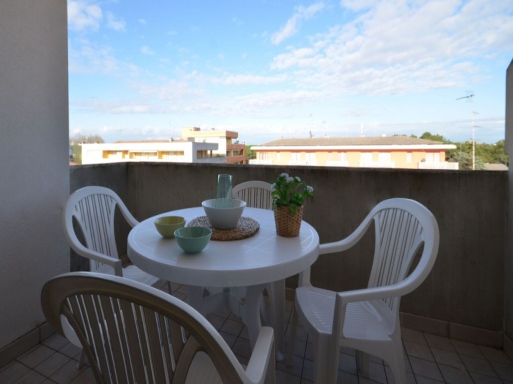 Un balcón con una mesa redonda y cuatro sillas blancas, una planta en maceta y vistas de edificios al fondo.