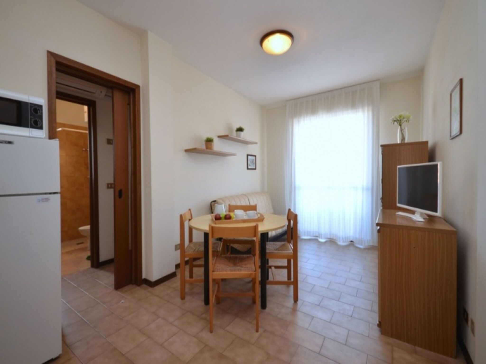 Een gezellig, compact leefruimte met kitchenette, een eethoek met een ronde tafel en stoelen, een televisie op een houten kast, en een raam met gordijnen.