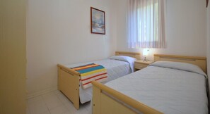 Een kleine, schone kamer met twee eenpersoonsbedden, een nachtkastje met lamp, een raam met gordijnen, en een ingelijst kunstwerk aan de muur.