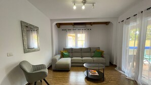 Ein Wohnzimmer mit Sofa, Sessel und Couchtisch. Ein Fenster mit Vorhängen und einer Deckenleuchte.