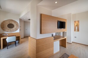 Ein modernes Hotelzimmer mit Schreibtisch aus Holz, einem Sessel, einem an der Wand montierten Fernseher und einem großen Schminkspiegel.