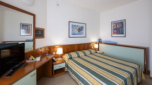 Ett hotellrum med en säng, ett skrivbord med en tv, en telefon och två inramade konstverk på väggen.