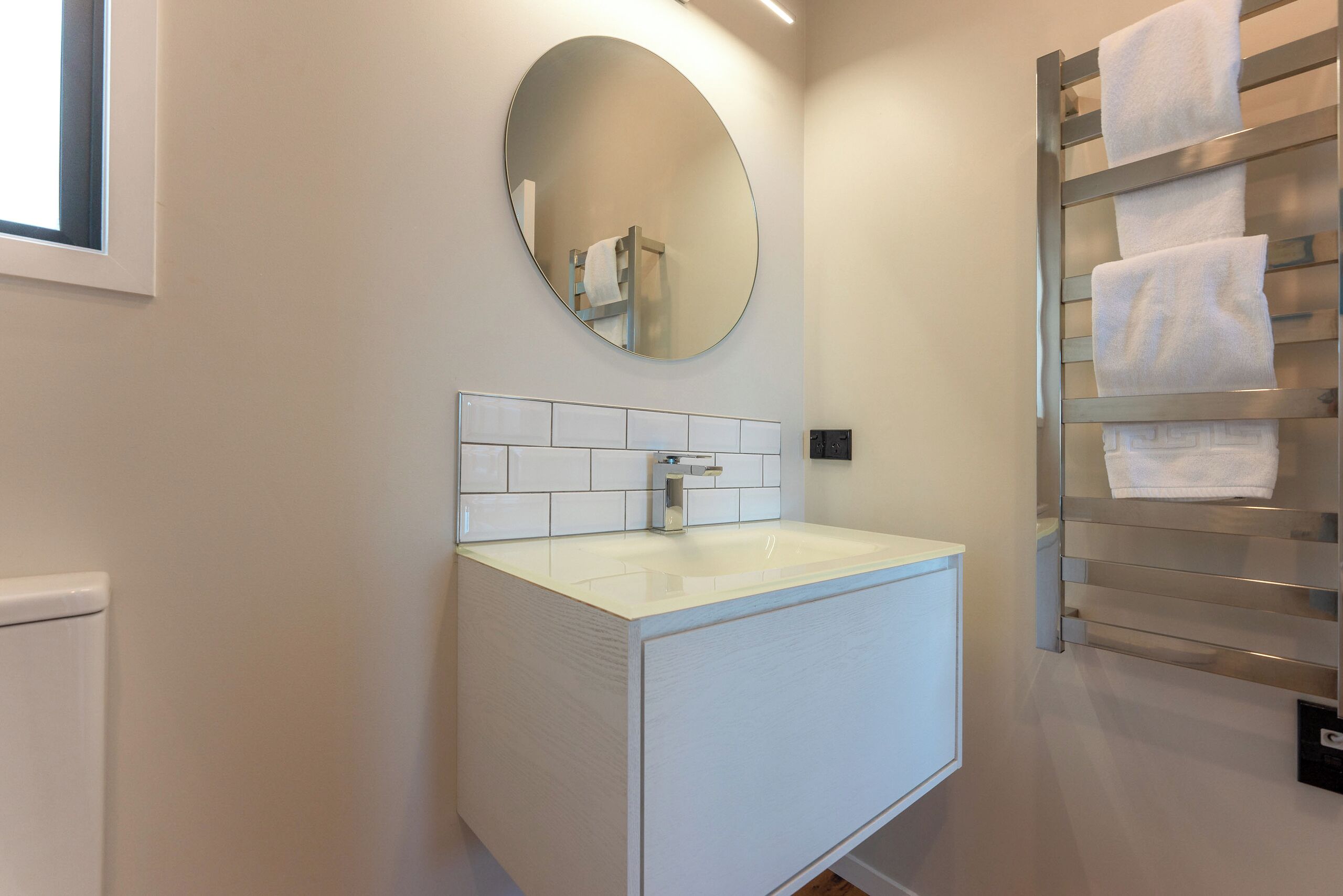 A modern bathroom with a white sink, a round mirror, and a towel rack with multiple towels.