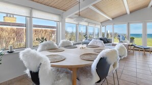 Un comedor amplio con una mesa redonda, sillas blancas de aspecto mullido y ventanas grandes que ofrecen vistas al mar.