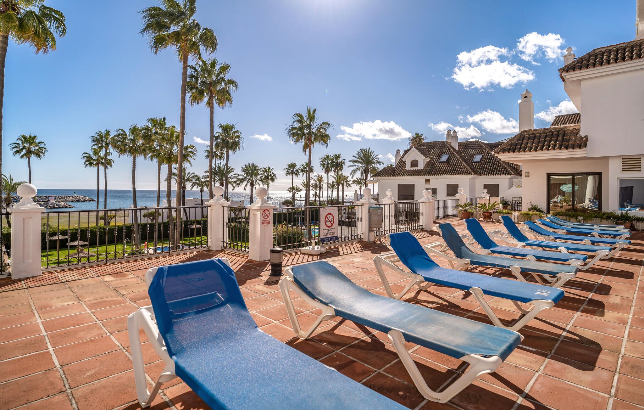 Un espace extérieur ensoleillé, aménagé avec des fauteuils bleus confortables, une piscine et une vue sur la mer.