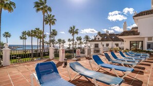 A sunlit outdoor seating area with blue lounge chairs, a pool, and a view of the sea.