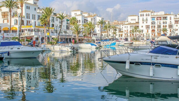 A marina with boats docked, surrounded by white buildings with balconies and palm trees.