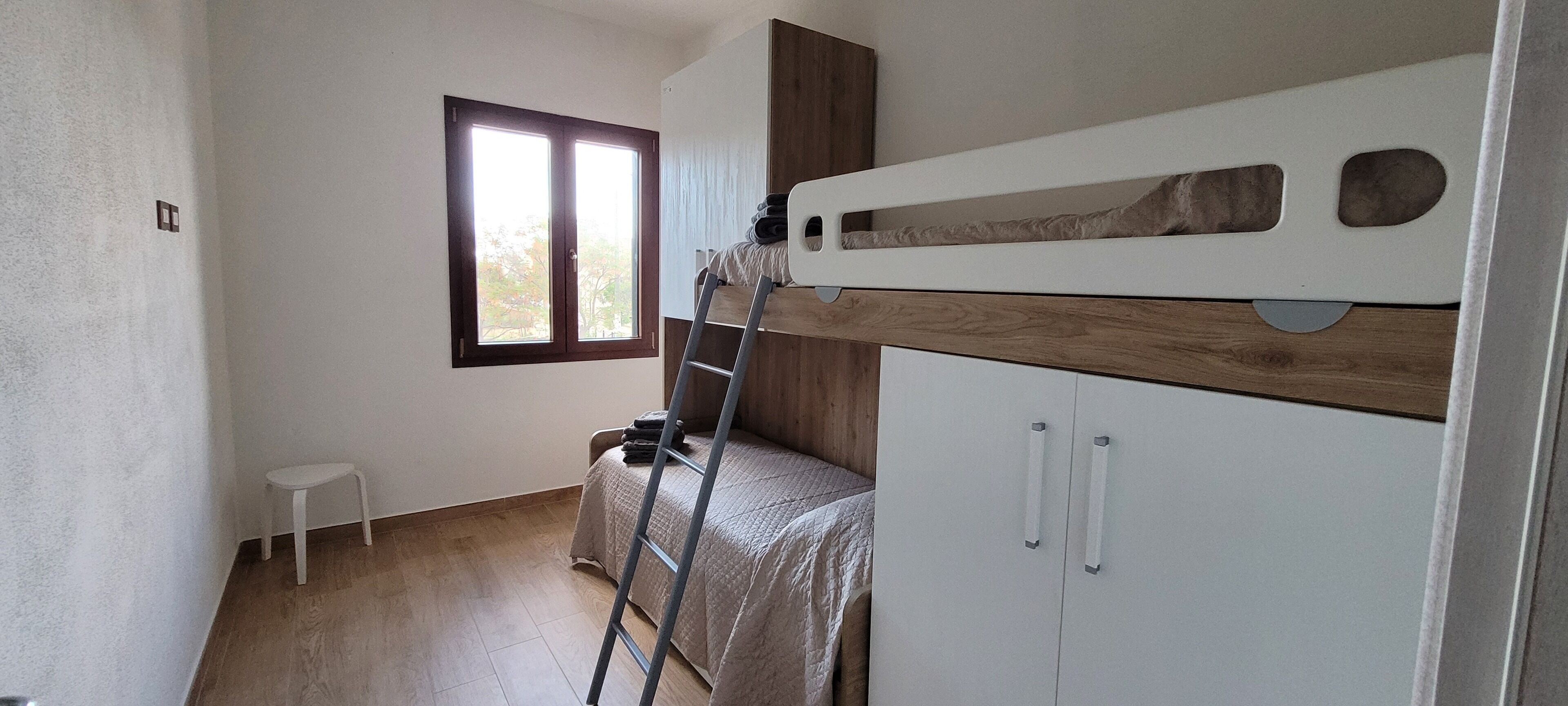 Una habitación con literas, un taburete pequeño blanco, una escalera y una ventana con vistas a árboles.