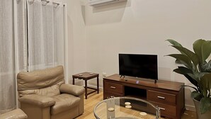 Een woonkamer met een beige bank, een glazen salontafel, een houten tv-meubel en een potplant.
