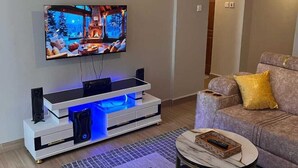 A modern living room with a flat-screen TV mounted on the wall, a white entertainment unit with blue LED lighting, a comfortable sofa with cushions, and a small round coffee table. - Martin Aviator Apartement (Kigali)