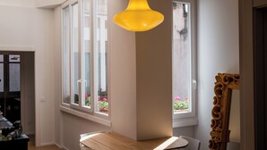 A room with a wooden dining table, a yellow pendant light, and a large window with potted plants.