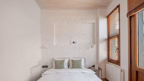 A bedroom with a white brick wall, a bed with white bedding, a wooden headboard, and a window with wooden blinds.
