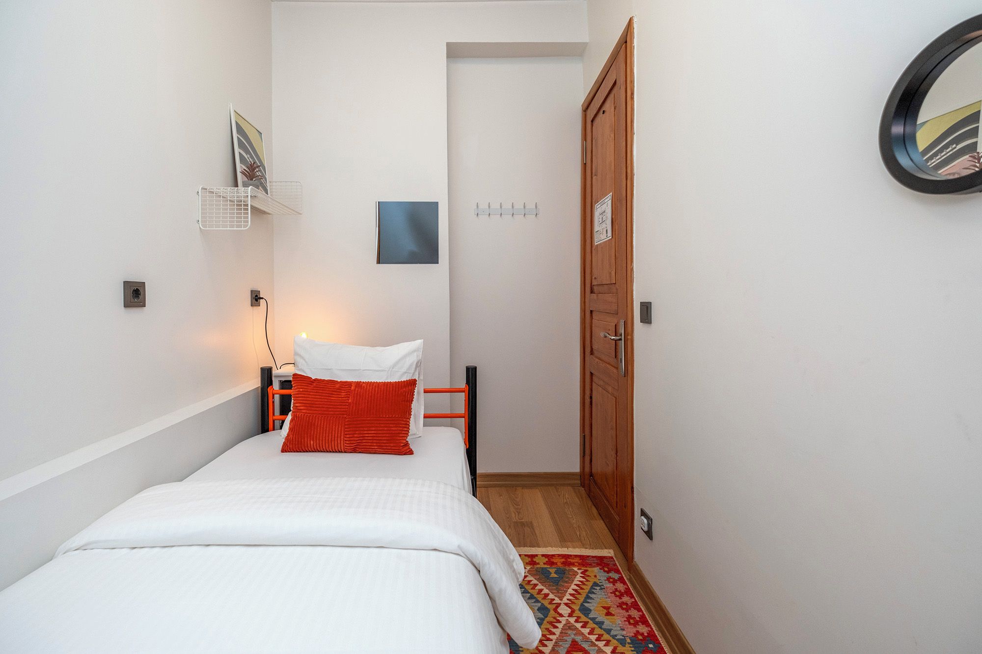 Une petite chambre à coucher, bien rangée, avec un lit, une table de chevet, une lampe, une porte et une étagère fixée au mur.