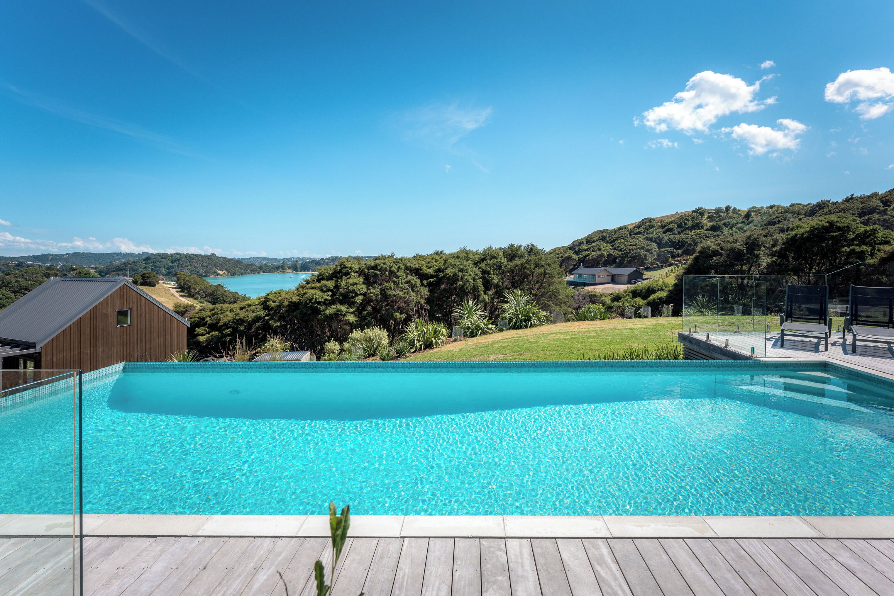 A clear blue swimming pool with a wooden deck in the foreground, surrounded by lush greenery and hills in the background.
