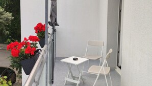 A small balcony with a white table and two chairs, a metal railing, and a potted plant with red flowers.