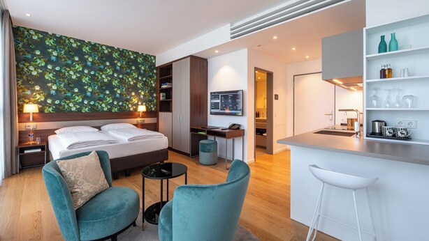 A modern hotel room with a large bed, two armchairs, a small round table, a kitchenette, and a TV.