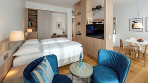 A modern hotel room with a bed, two blue armchairs, a small round table, a TV, and a dining area with a table and chairs.