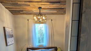 Un dormitorio con techo de madera, una cama con cabecera, una mesita de noche con lámpara, una ventana con cortinas y un cuadro enmarcado en la pared.
