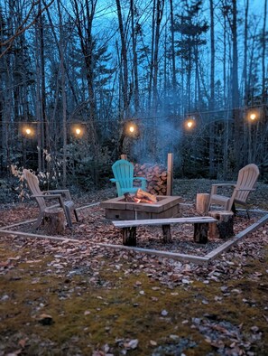 Uma área externa arborizada com assentos Adirondack, uma fogueira e luzes de corda.