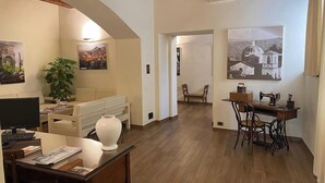A spacious living room with a wooden floor, a white sofa, a coffee table, and a desk with a sewing machine. There are two paintings on the walls and a potted plant. - Borgo Ospitale (Rotonda)
